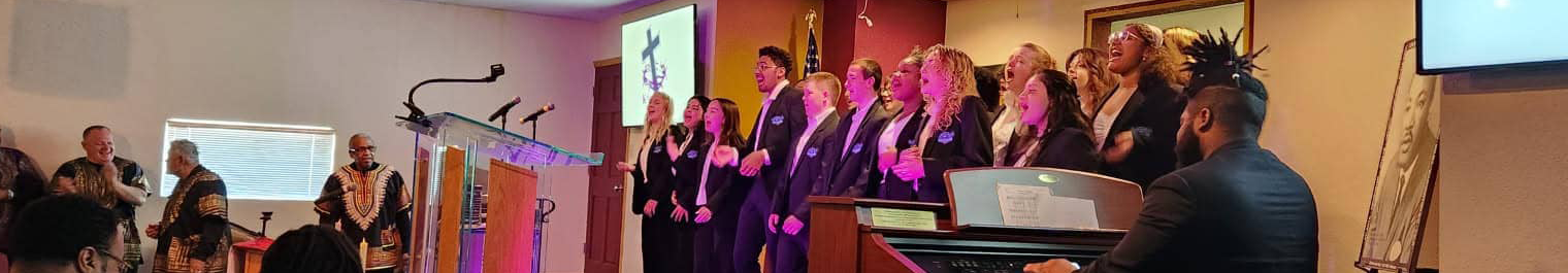Mixed choir performs on a church stage with a pianist at a keyboard to the right; audience on the left and a cross image on a screen behind them.