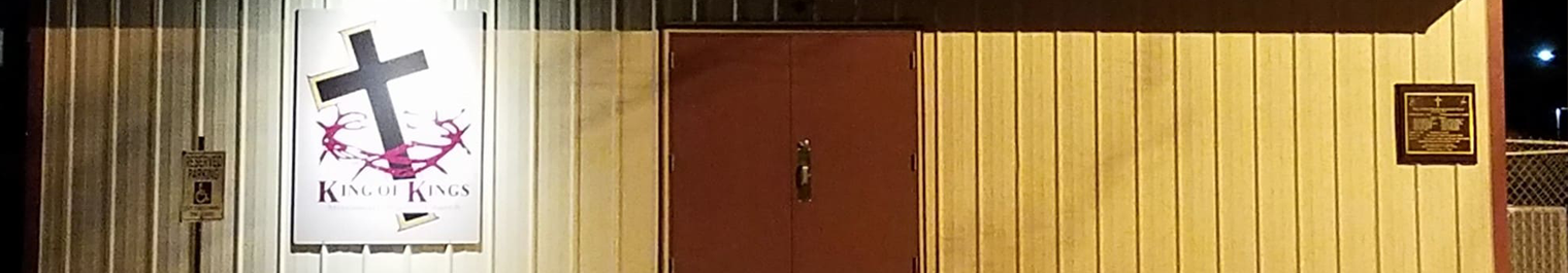 Night exterior of a metal-siding building with a large 'King of Kings' sign featuring a cross, brown central doors, and plaques on the walls.